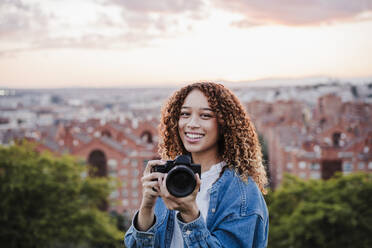 Smiling woman holding camera during sunset - EBBF03663