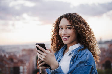 Happy woman holding smart phone in front of sky - EBBF03668