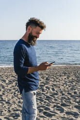 Lächelnder Mann, der am Strand stehend ein Mobiltelefon benutzt - MGRF00228