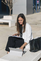 Mature businesswoman with book using digital tablet on sunny day - JRVF00668