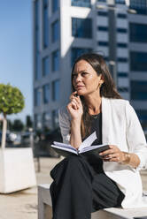 Weibliche Unternehmerin schaut weg, während sie an einem sonnigen Tag mit einem Buch sitzt - JRVF00670