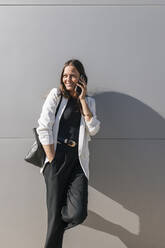 Smiling businesswoman with hand in pocket talking on mobile phone in front of wall - JRVF00671