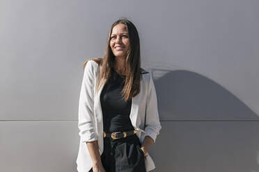 Smiling mature businesswoman standing in front of gray wall - JRVF00672