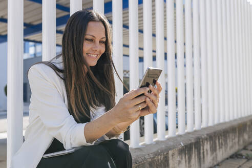 Eine Unternehmerin benutzt ein Mobiltelefon, während sie vor einem Zaun sitzt - JRVF00690