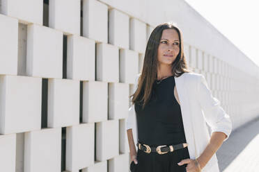 Mature businesswoman standing with hands in pockets by wall on sunny day - JRVF00693