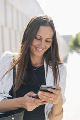 Geschäftsfrau lächelt bei der Benutzung eines Mobiltelefons an einem sonnigen Tag - JRVF00695