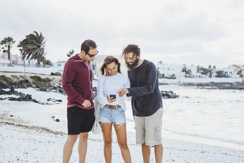 Freunde, die am Strand stehen und ein Mobiltelefon benutzen - DGOF02185