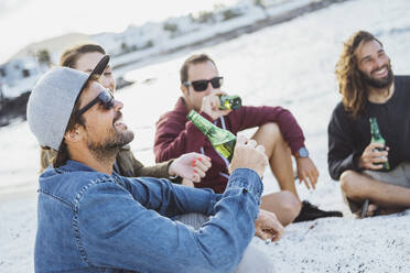 Lächelnder Mann mit Getränk, der mit Freunden am Strand sitzt - DGOF02189