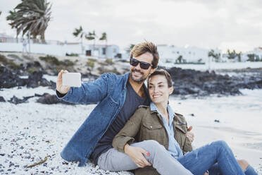 Smiling man taking selfie with woman while sitting at beach - DGOF02197