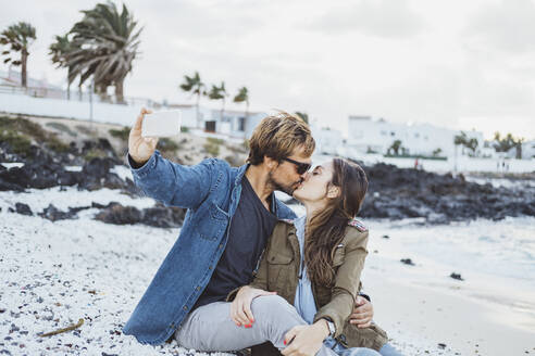 Freund küsst Freundin, während er ein Selfie mit dem Smartphone am Strand macht - DGOF02198