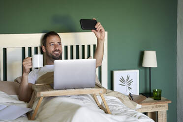 Lächelnder Mann mit Kaffeetasse nimmt Selfie durch Smartphone im Schlafzimmer - VEGF04534