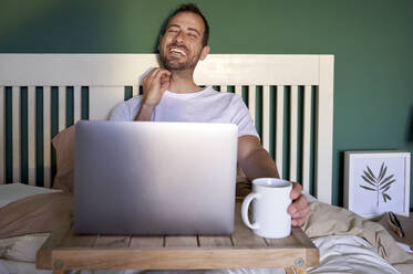 Lächelnder Mann sitzt mit Laptop und Kaffeetasse auf dem Tisch im Bett - VEGF04547