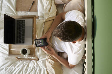Ein Mann führt einen Videochat mit einem Freund über sein Mobiltelefon, während er im Bett am Laptop sitzt - VEGF04548