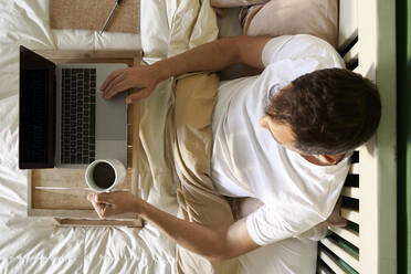 Mann sitzt mit Kaffeetasse und Laptop auf dem Tisch im Bett - VEGF04549