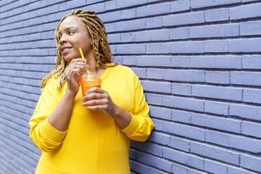 Smiling woman holding juice in disposable cup leaning on brick wall while looking away - IFRF00710