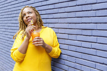 Thoughtful woman holding juice looking up while leaning on brick wall - IFRF00711