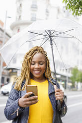 Lächelnde Frau, die ein Smartphone benutzt und einen Regenschirm hält - IFRF00713