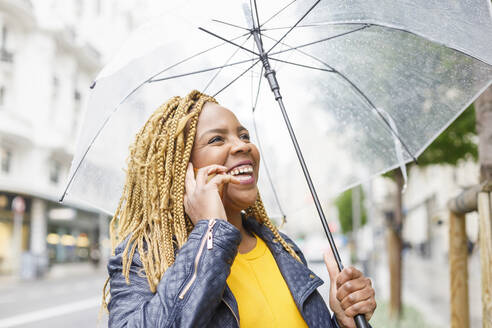 Fröhliche Frau mit geflochtenem Haar telefoniert und hält einen Regenschirm - IFRF00714