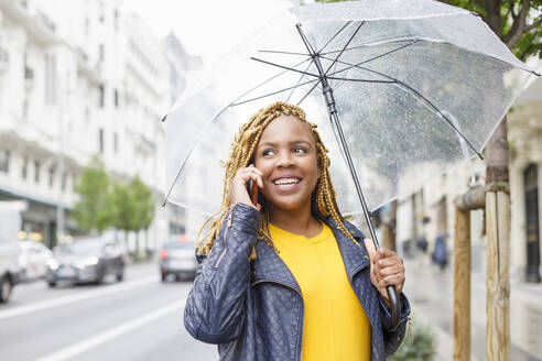 Lächelnde Frau mit geflochtenem Haar, die mit einem Regenschirm telefoniert - IFRF00715