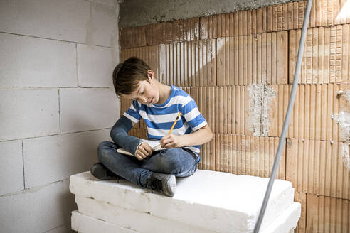 Boy with broken arm writing in book while sitting on block during renovation - HMEF01222