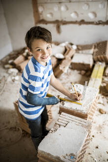 Smiling boy measuring brick while standing at house during renovation - HMEF01227