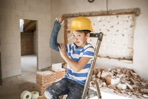 Boy flexing broken arm muscles while sitting on ladder during house renovation - HMEF01238