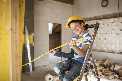 Smiling boy looking at pocker rule while sitting on ladder at construction site - HMEF01239