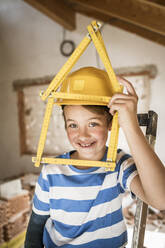 Junge lächelt, während er auf dem Dachboden einen hausförmigen Zollstock hält - HMEF01243
