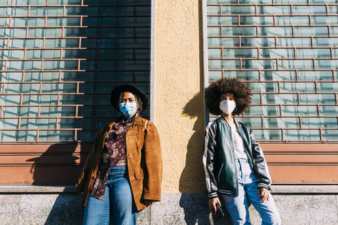 Young women wearing protective face masks standing with social distance in front of building - MEUF02882