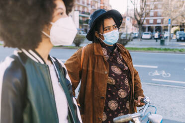 Young woman wearing protective face mask walking with bicycle and friend on road - MEUF02883