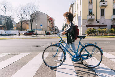 Young woman wearing protective face mask crossing road with bicycle on sunny day - MEUF02888