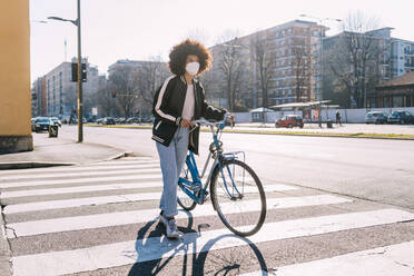 Woman wearing protective face mask walking with bicycle on road in city - MEUF02894