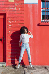 Cheerful woman jumping in front of red building during sunny day - MEUF02908