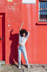 Woman standing with hand raised in front of red building - MEUF02917