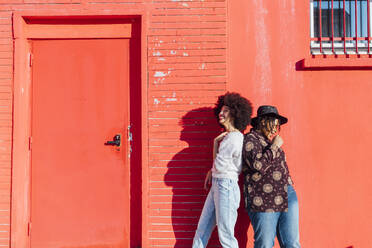 Lächelnde Freundinnen stehen Rücken an Rücken vor einem roten Gebäude - MEUF02959