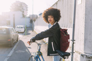 Junge Afro-Frau mit Rucksack und Fahrrad auf dem Fußweg stehend - MEUF02985