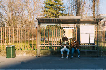 Female friends with face mask using mobile phone at bus station - MEUF02995