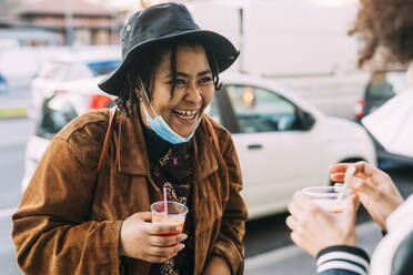 Young woman with face mask and drink smiling while looking at friend - MEUF02996