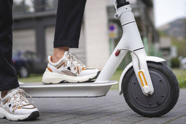 Businesswoman wearing sports shoe standing with electric push scooter on footpath - SNF01443