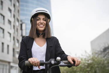 Businesswoman wearing cycling helmet smiling while standing with electric push scooter - SNF01446