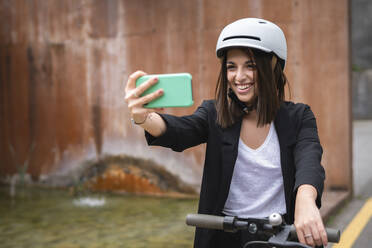 Lächelnde Geschäftsfrau mit Elektroroller und Fahrradhelm, die ein Selfie mit ihrem Smartphone macht - SNF01465