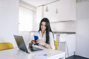 Young beautiful woman using mobile phone while sitting at home office - DGOF02220