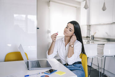 Woman wearing in-ear headphones while sitting at home office - DGOF02226