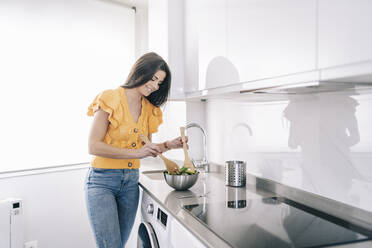 Smiling woman making salad while standing at kitchen counter - DGOF02229