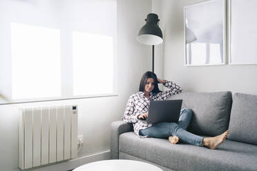 Junge Frau benutzt einen Laptop, während sie sich zu Hause auf dem Sofa ausruht - DGOF02238