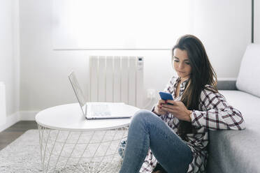Junge Frau mit Laptop, die ein Mobiltelefon benutzt, während sie im Wohnzimmer sitzt - DGOF02245