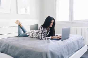 Junge Frau, die auf dem Bett liegend einen Laptop benutzt - DGOF02251