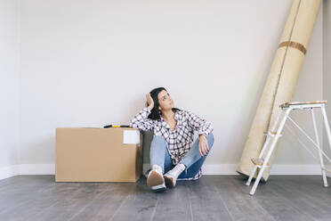 Thoughtful woman sitting by box on floor at home - DGOF02256