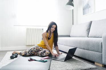 Junge Frau benutzt einen Laptop, während sie auf einer Übungsmatte im Wohnzimmer sitzt - DGOF02259