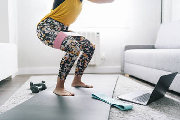 Woman with resistance band crouching while learning through laptop at home - DGOF02261